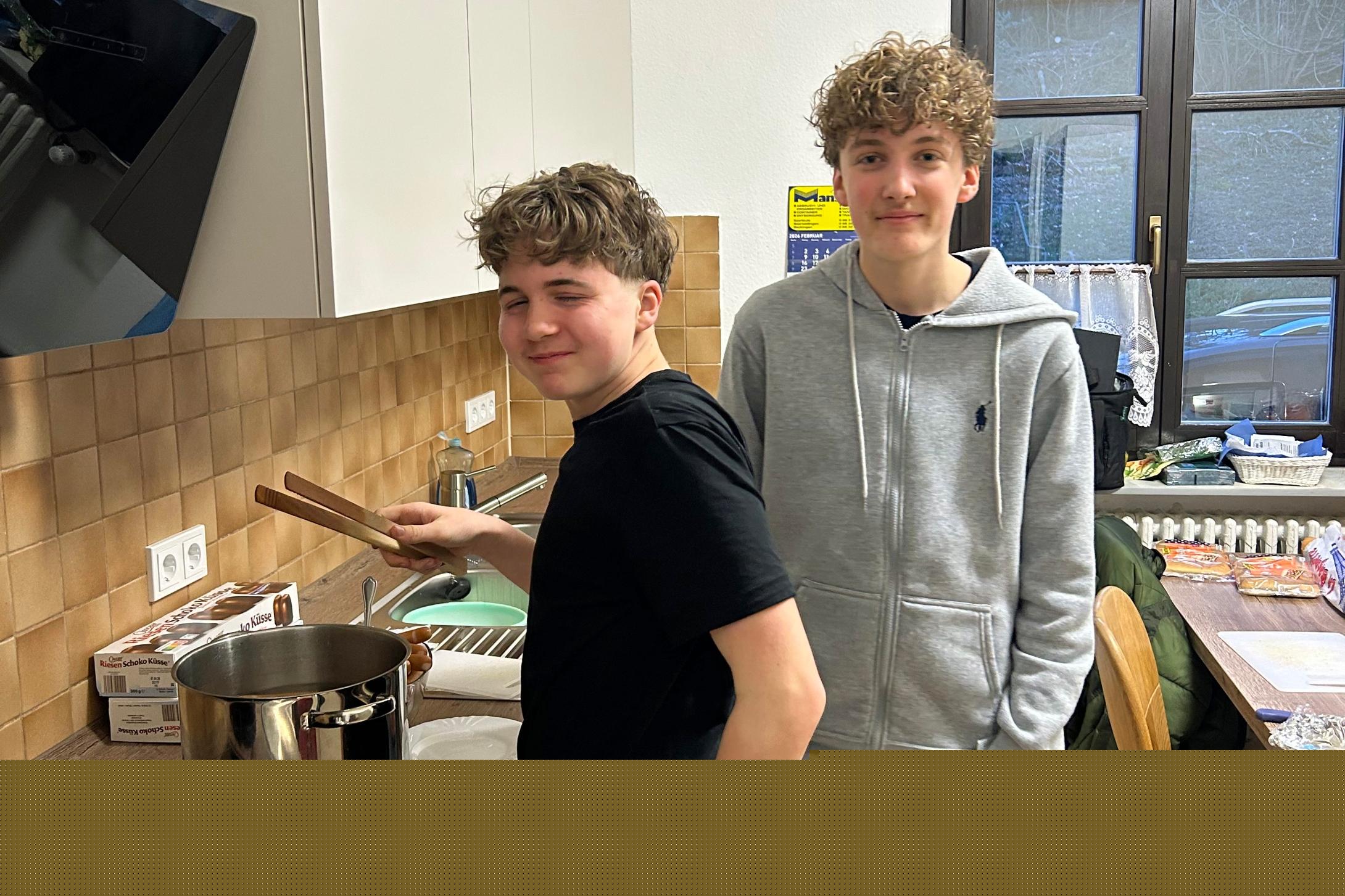 Zwei Jugendliche stehen in einer Küche. Eine Person steht am Herd und rührt mit Küchenzangen in einem großen Edelstahlkochtopf auf der Arbeitsplatte. Daneben liegen ein Topfdeckel und Zutaten wie Hotdog-Brötchen. Die zweite Person steht leicht seitlich dahinter, mit den Händen in den Hosentaschen. Im Hintergrund sind Küchenschränke, ein Fenster, ein Esstisch mit Stühlen und weitere Lebensmittel zu sehen.